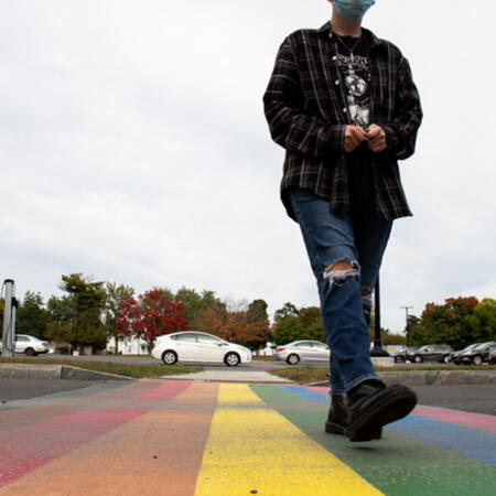 A photograph of a student walking towards the camera, along a rainbow colored pride crosswalk.