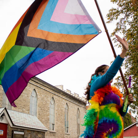 A photograph of a Pride Parade participant marching and flying the inclusive rainbow pride flag with their arm outstretched.