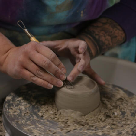 A dimly lit photograph of a tattooed artist carving into a clay slab on the wheel.