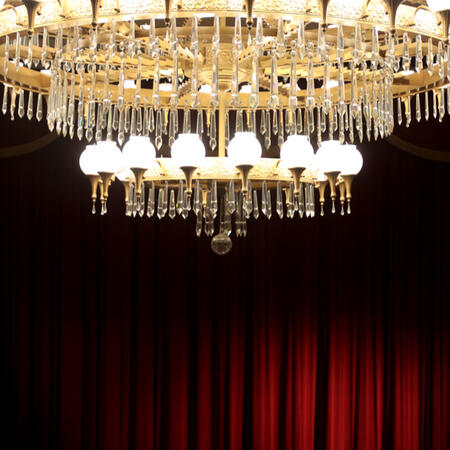 A photograph of a grand gold crystal chandelier against a red curtain background with a spotlight.