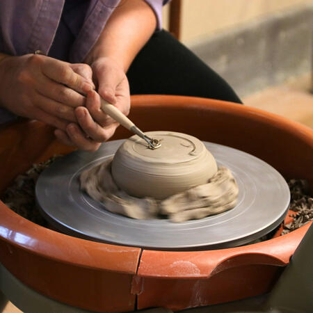 A brightly lit photograph of an artist's hands carving into a clay slab as the wheel spins.